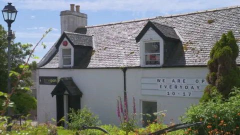 A small white-walled cottage behind an area of shrubbery. 