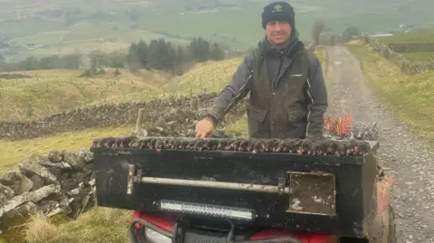 Mikey Fullerton Mikey Fullerton is a man in his 40s wearing a green barber-type jacket and green woolly hat. 