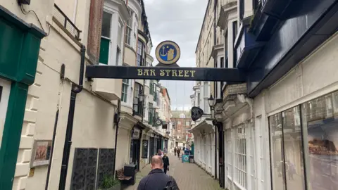 A narrow pedestrian street lined with older-style buildings on both sides. At the top of the scene is a black overhead sign with gold text that reads:
'Bar Street'.