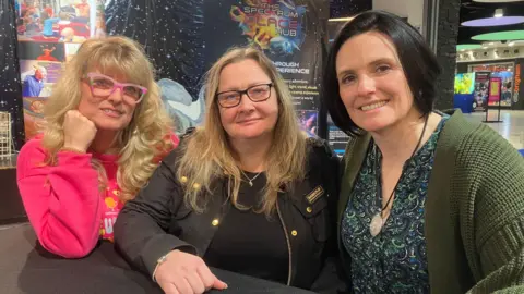 Three women are seated together at a table inside a science‑themed venue, with colourful displays and signage in the background. One wears a bright pink top, another a dark jacket, and the third a patterned top with a green cardigan.