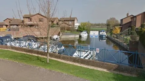 Riverside Mead, Peterborough, showing a piece of water, a bridge, boats on the water and houses either side of the river. There is a tree by the bridge. 