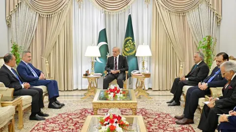 Turkish Foreign Ministry / Handout/Anadolu via Getty Images Men wearing suits sitting in a room of gold armchairs. The Pakistani flag is standing in the background.