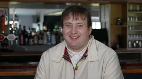 SHAUN WHITMORE/BBC Macaulay Robinson sitting in the clubhouse at Kirkley and Pakefield Football Club in Lowestoft in front of the old wooden style bar. He is wearing a beige scooter style jacket with a poppy on it and a tartan lining. He is wearing a white polo shirt and has a brown fringe. He has a wide smile and is of stocky build.