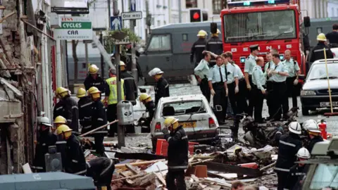 PA The fire service and police officers inspecting the damage caused by the Omagh bomb. There is a white car among them with wood, metal and other materials scattered across the road by the explosion.