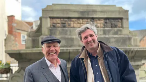 BBC Two men smiling and looking at the camera. On the left is Nick Cragg, a man wearing a grey flat cap, a grey suit over a pink button up shirt. On the right is James Capper, he's wearing a number of jackets, on top is a blue one. He has long grey hair. 