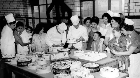 Getty Images A black-and-white image of a table laden with about 20 Chistmas cakes. Three men in chef's whites and hats iced the cakes, watched by a group of women. Soe of the women are in nurses' uniforms and hats, others are in normal clothes, and one is in pyjamas. Two children in night clothes are being held by the nurses while another stands alongside, looking on.