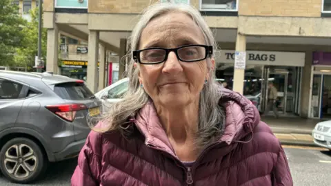 A woman with a purple coat, black glasses and long grey hair. She is stood in front of shops and cars. 