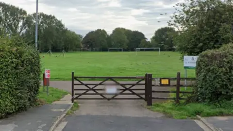 Google shot of an entrance to St Aiden's Park. There is a large wooden gate and football goals in the distance. There is also a red bin to the side of the gate.