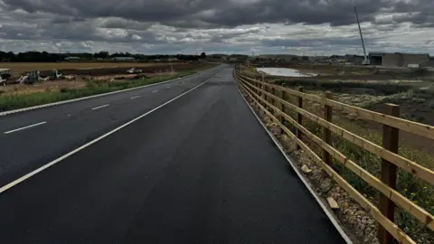 National Highways A new section of a new road, showing freshly laid tarmac, a wooden fence, along the righthand side, white markings on the road and machinery in the distance, including a large crane.