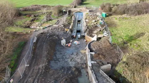 Drone view of a construction site for a canal lock surrounded by muddy ground and temporary fencing, with green fields in the background.