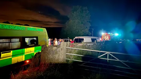 The scene on the road near the cave entrance, at night, with an emergency ambulance in the foreground