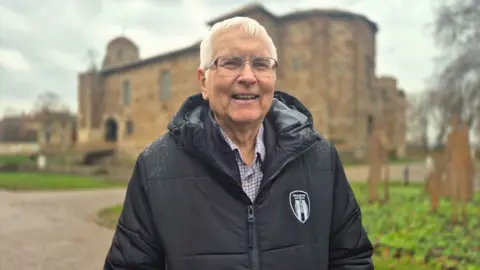 Stuart Woodward/BBC Sir Bob Russell stands in front of Colchester Castle. He is wearing a black hooded jacket with a Colchester United logo on his left chest, and a chequered shirt underneath. Sir Bob has white hair and spectacles, and is smiling at the camera