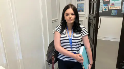 A girl wearing a white t-shirt with black stripes on it stands holding her bag and a folder. A door can be seen open behind her, with posters and leaflets on the wall of the corridor.