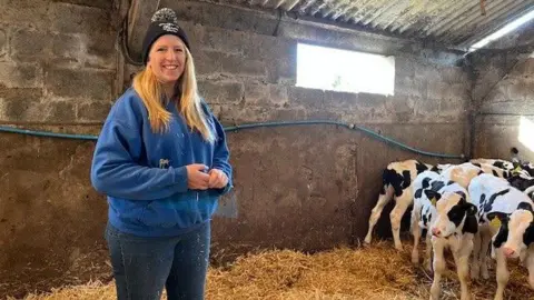 Woman with long blonde hair dressed in blue hooded top and wearing a black woolly hat stood in barn alongside young cows