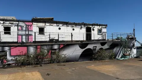 HMS President with grey and pink paintwork. The boat looks dilapidated and lots of weeds are growing up its sides.