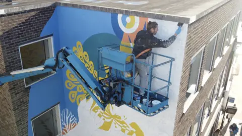 Jamie Niblock/BBC A drone image of Atma the artist standing on a cherry picker that has been raised to the top floor of a brick building. He paints on its outer corner. He has a harness around his torso that is attached to the cherry picker. 