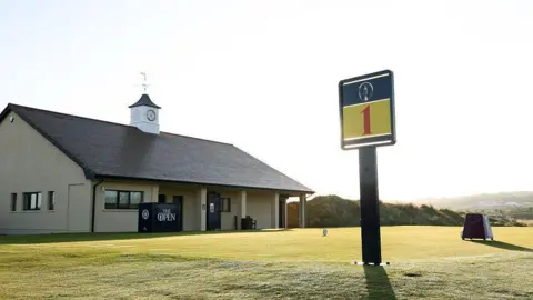 Getty Images A large sign has the number '1' on it in red font. It's sits on a green grass tee box. Beside the tee box is a small cream coloured building. The building has a white clock on top of it.