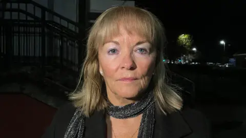 A woman with shoulder length blonde hair, looking at the camera. She is wearing a black sparkly scarf and a black jacket. It is dark outside.