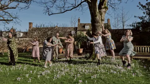 Dancers from the Remarkable Dance Company performing outside in a garden among blooming purple crocus flowers. There are a few benches throughout the garden.