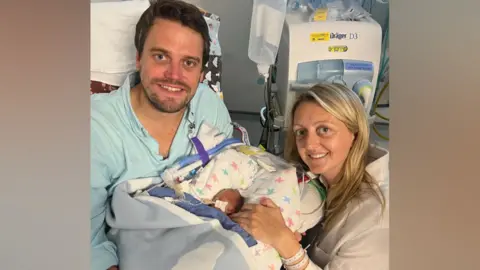 Nick and Libby Powell A couple smile as they pose with their premature son. The man, who has short brown hair and is sitting down in a chair, is holding the baby while his wife, who has long blonde hair, is crouching down beside them with her hand resting near the baby's head.