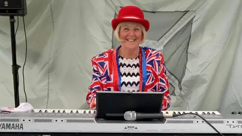 A woman in a Union Jack blazer and zig zag patterned black and white smiles while sitting at a mini piano. She has short blonde hair in a bog and is wearing a red bowler hat. She is in a tent, with grey material behind her.