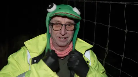 A man wearing a felt hat in the shape of a frog and dark-rimmed glasses, he has a hi-vis jacket and black leather gloves. 