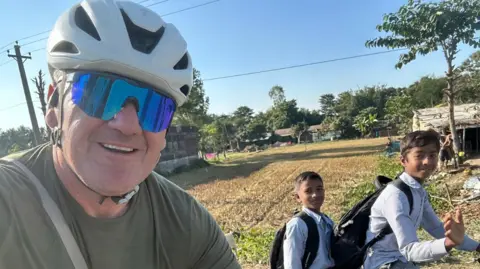 Stephen Aslin Stephen, wearing a helmet, sunglasses and a green t-shirt, on his bike in a rural area in Asia - two children cycling past smile and wave at the camera.