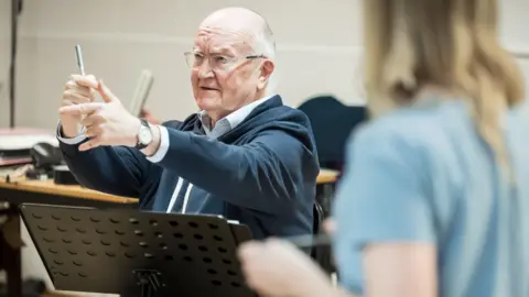 York Theatre Royal/Marc Brenner John Doyle sits at the front of a rehearsal room, gesturing as he leads a rehearsal.