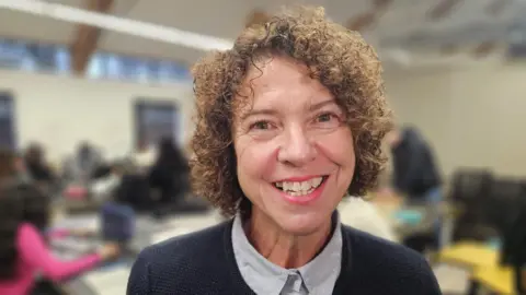 Sam Wilson has short curly light brown hair, she is smiling at the camera and is wearing a light blue shirt with a navy cardigan.