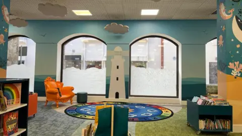 Inside Jersey's Children's Library. It is nautical themed, there is a lighthouse and sea creatures painted on the walls. An orange arm chair is in the back corner, and the bookshelves are shaped like boats. There is a mat on the floor with the letters of the alphabet around the outside. The windows of the library have been frosted to look like waves.