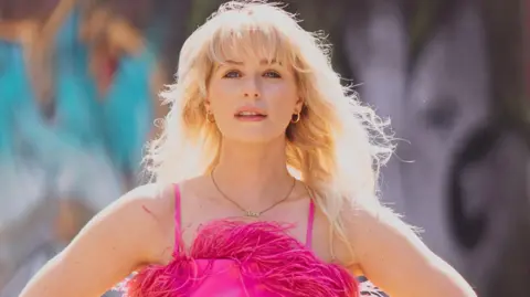 Jess Davies wears a neon pink jumpsuit, with feather fringe detailing. She's wearing a gold necklace and hoop earrings, with her wavy blonde hair swept in the wind.