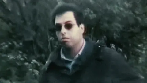 Danilo Restivo, a man with bushy dark hair, wearing dark glasses and a dark coat over a light collared shirt, pictured in a slightly blurred snatched image in front of some bushes or trees.