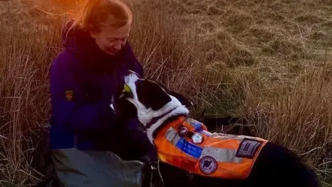 A woman wearing waterproofs playing with a collie dog wearing an orange fluorescent jacket in a field, with the sun setting behind them