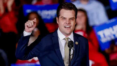 Shutterstock Matt Gaetz stands with one arm raised in the air, speaking before a crowd of people holding red and blue signs. He has on a blue suit and gold tie