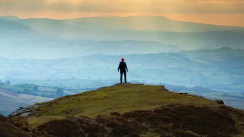 A person is standing on top of a mountain during sunset looking down on roaming fields.