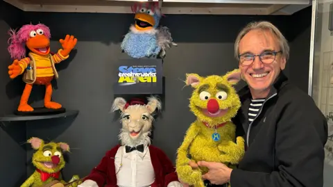 Steve Allen is holding a green puppet which is reminiscent of the Muppets characters. There are four other puppets on static display behind him, including one of the characters from Fraggle Rock.