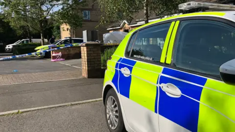 Police cars near the scene of a house in Gendros, Swansea, with police tape and a child's toy in the background 