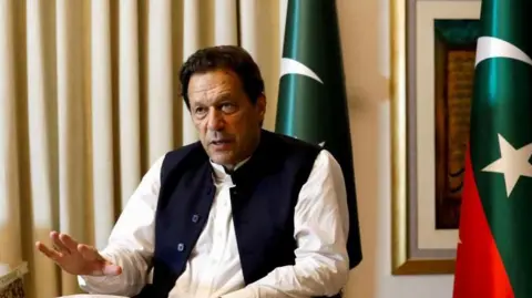 Former Pakistani Prime Minister Imran Khan speaks with Reuters during an interview, in Lahore, Pakistan March 17, 2023. He can be seen seated with his hand outstretched and speaking. Two Pakistani flags are behind him