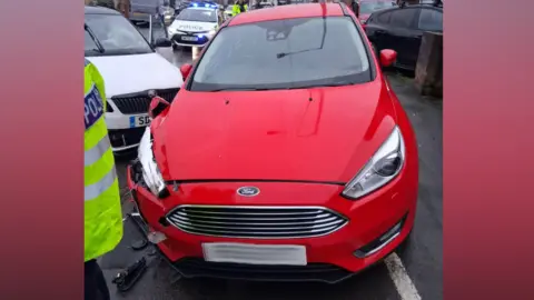 Hugh Reynolds A red Ford Focus with significant damage to its front left corner is parked on a residential street. A white car with damage to its front is visible behind it, along with a police car with flashing lights. A person in a high-visibility jacket stands to the left of the frame.
