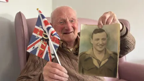 An old, bald, man wearing a brown knit jumper and sat in a pink armchair, smiling at the camera. He is holding up a Union Jack flag and a photograph of him as a young man with short brown hair, wearing a soldier's uniform.