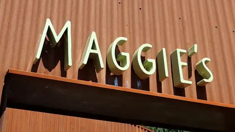 The sign of a Maggie's Centre. The word is in a light green colour on an orange corrugated metal background.