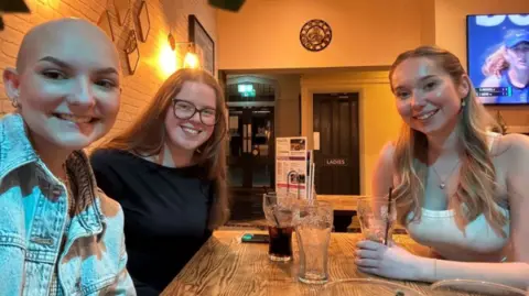 Lilli Hales Three women with the one on the left who has a bald hairstyle and is wearing a denim jacket and smiling. Next to her is a girl with long brown hair, large glasses and a black top. To the right is another woman sat across the table holding a cup. She has long blonde hair and is wearing a white vest.