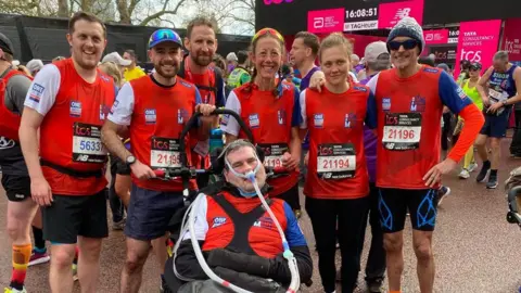 Stand Against MND Group stood at the finish of a sporting event, with six people in matching red tops stood with Sam Perkins, who is in a wheelchair.