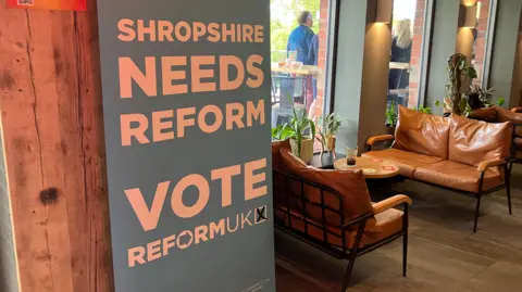 Blue pull-up banner, which says "Shropshire news Reform; Vote Reform UK" against a wooden wall in a bar with leather sofas and round tables