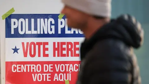 A man in a beige tuque and a black puffer jacket walks by a red, white and blue sign that reads in English and in Spanish 'Polling place, Vote here". 