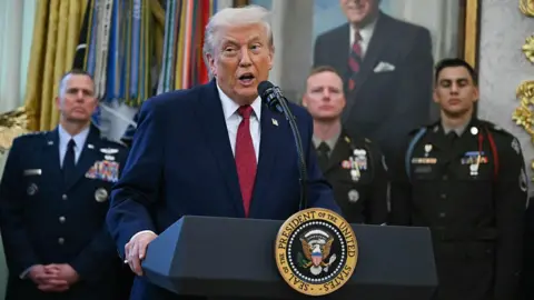 Getty Images US President Donald Trump wearing a blue suit and red tie in the Oval Office in front of US servicemembers on 15 December. 