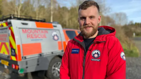 Aaron Duke, wearing a red jumpsuit in a mountain rescue uniform, stands in front of a 4x4 rescue vehicle. He smiles looking into the room. 