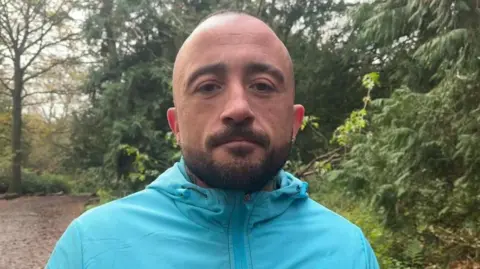 Ellen Knight/BBC A man with cropped dark hair is pictured looking into the camera with a serious expression. He is wearing a bright blue raincoat. He is standing in woodland, in front of a cluster of evergreen trees. Behind him, a path stretches out into the distance, covered in fallen leaves. The sky is grey and overcast. 