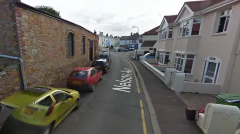 Nelson Avenue has a beige house on the right hand side and a brick building on the left hand side of the road. Several cars like the left hand pavement and there is a single yellow line along the right of the road.