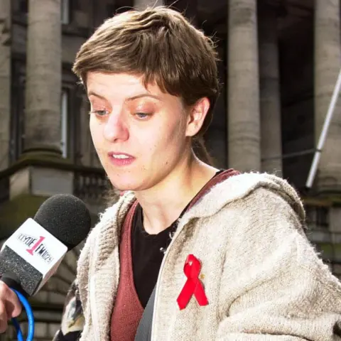 PA Media Sandy Brindley in a photo taken in 2000. She has short, cropped hair and someone is holding a microphone up to her. She is wearing a cream cardigan over a black and brown top. She has a red Aids ribbon pinned to her